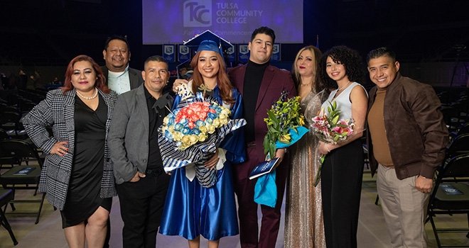 TCC Graduate Abigail Molina wears a blue graduation cap and gown while she holds a bouquet of flowers flanked by her family.