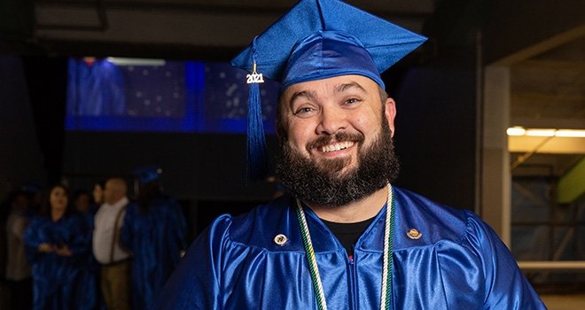 Smiling TCC Graduate with beard, wears blue cap and gown.