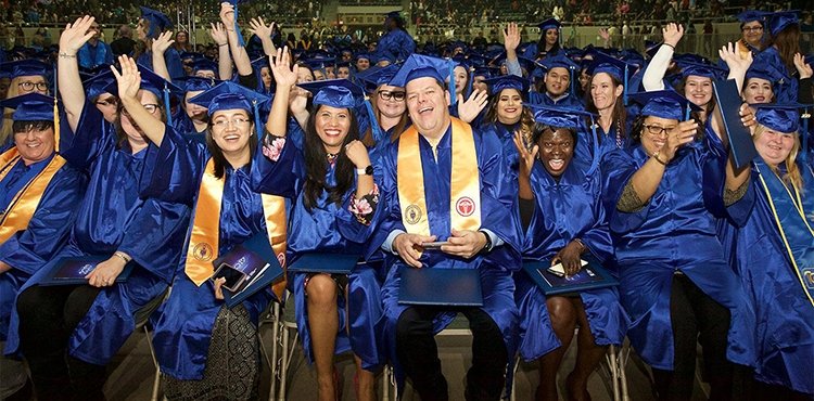 Seated TCC graduates wear caps and gowns and smile and wave at the camera.