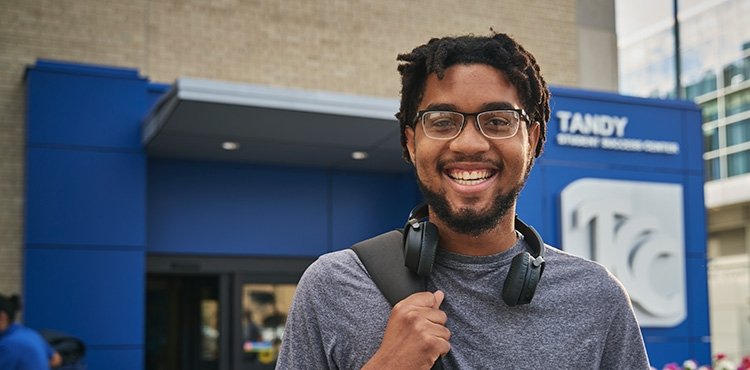 Smiling young black man stands with back pack over one shoulder outside Tandy Student Success Center.