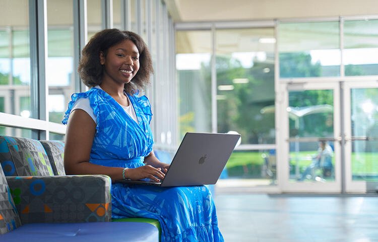 A smiling, female TCC student sits on a couch with her laptop.